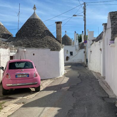 Trulli bianchi con tetti a cono lungo un vicolo ad Alberobello, con una Fiat 500 rosa parcheggiata accanto alle case storiche