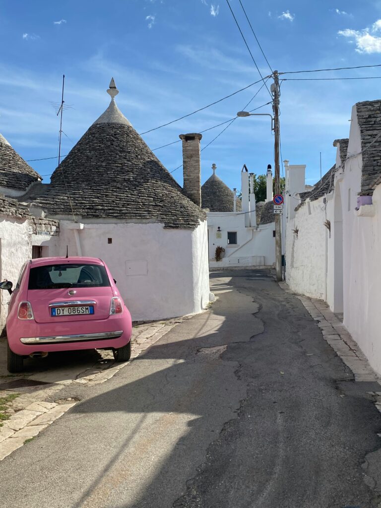 Trulli bianchi con tetti a cono lungo un vicolo ad Alberobello, con una Fiat 500 rosa parcheggiata accanto alle case storiche