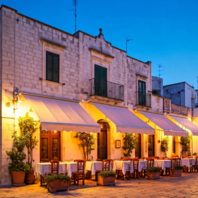 Facciata in pietra con tavolini all’aperto illuminati da luci calde in un centro storico pugliese al tramonto