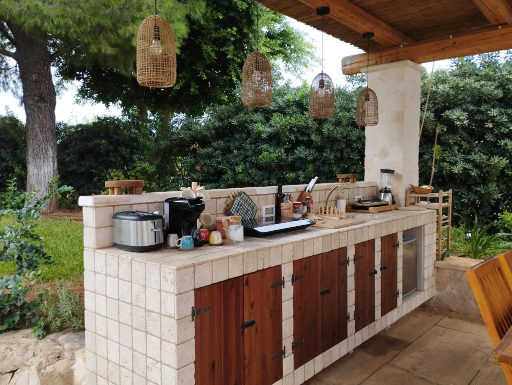 Cucina esterna in pietra della Villa Agapanthus con pensili in legno, lampade in vimini e vista sul giardino mediterraneo