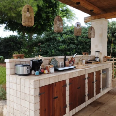 Cucina esterna in pietra della Villa Agapanthus con pensili in legno, lampade in vimini e vista sul giardino mediterraneo