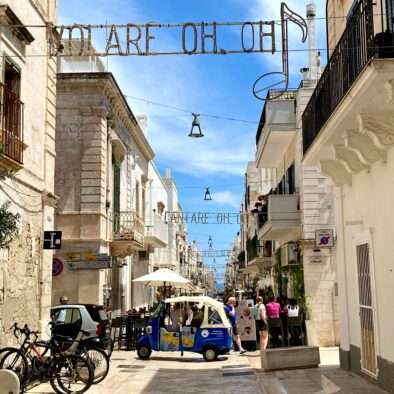 Centro storico di Polignano a Mare con la scritta "Volare oh oh", omaggio a Domenico Modugno, tra edifici bianchi e ape car