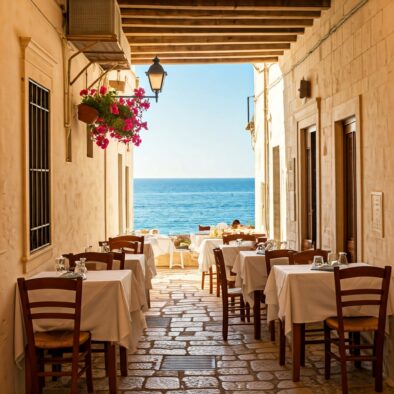 Vicolo con tavoli all’aperto apparecchiati e vista sul mare in un borgo pugliese, tra muri bianchi e fiori colorati